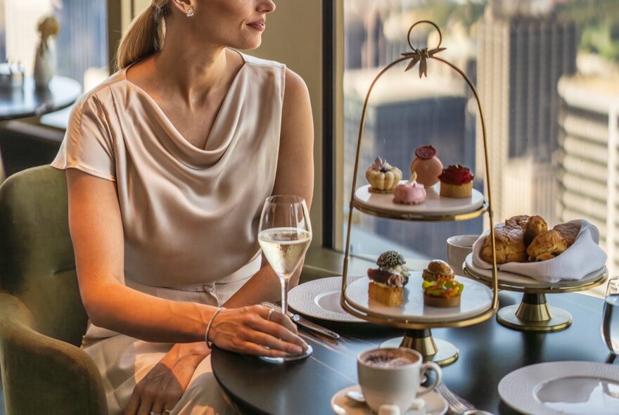 A woman in a softly draping silk dress holding a glass of wine in her hand, seated at a table that is set for a high tea with small morsels of sweet and savoury food on a two-tiered plate. 