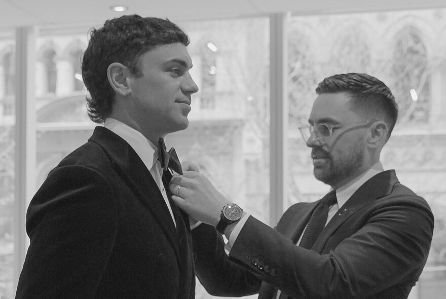 A sharply-dressed tailor putting the finishing touches on a young man's suit.