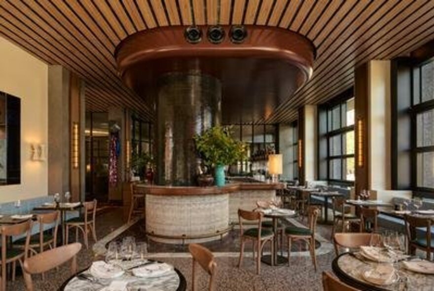 Interior view of a modern restaurant with a central circular bar, wooden ceiling beams, and set dining tables.