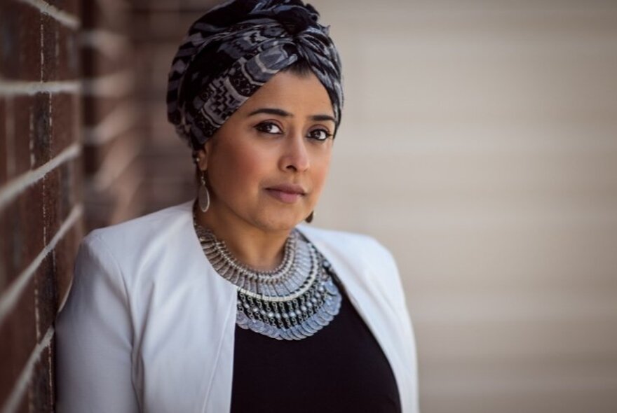 Tasneem Chopra leaning against a wall looking serious with a scarf covering her hair and a beaded collar.