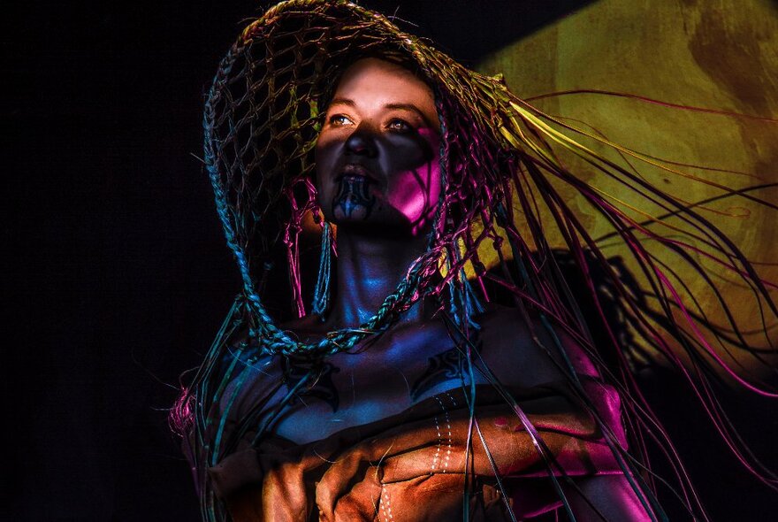 A person wearing a woven basketry headdress, dark face makeup, and looking up into the distance.