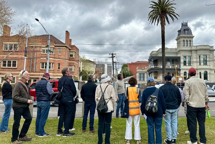 A tour guide leading a walking street tour, pointing to several Victorian-era buildings in the background, with a few people standing around listening on a small nature reserve.