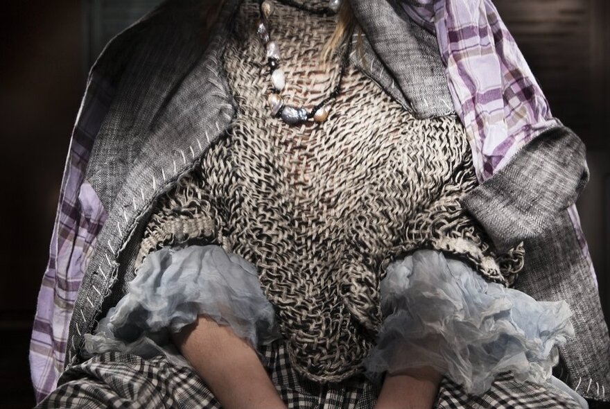 Close up of a person draped in a large looped knitted jumper and a purple checked jacket. 