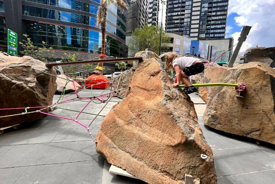 Child clambering over large rocks in a city playground space with skyscrapers visible in the background.