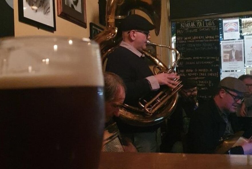 A man wearing a newsboy cap, playing a brass instrument in a pub, a glass of Guinness  on a table in the foreground.