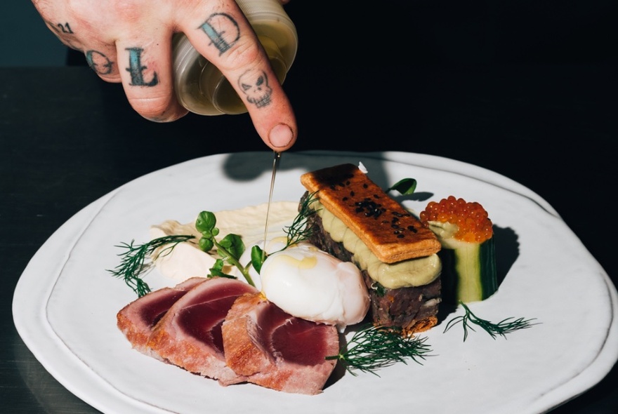 Tattooed hand adding a dressing to a plate loaded with ham, eggs and side dishes.