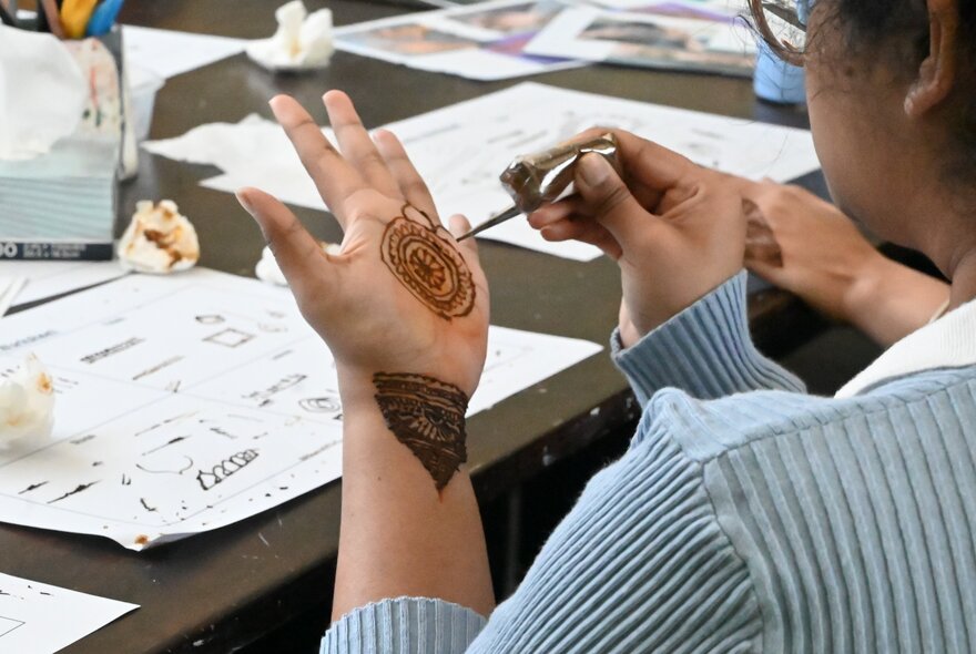 A person applying henna art to their hand using a fine applicator cone as part of a henna art workshop.