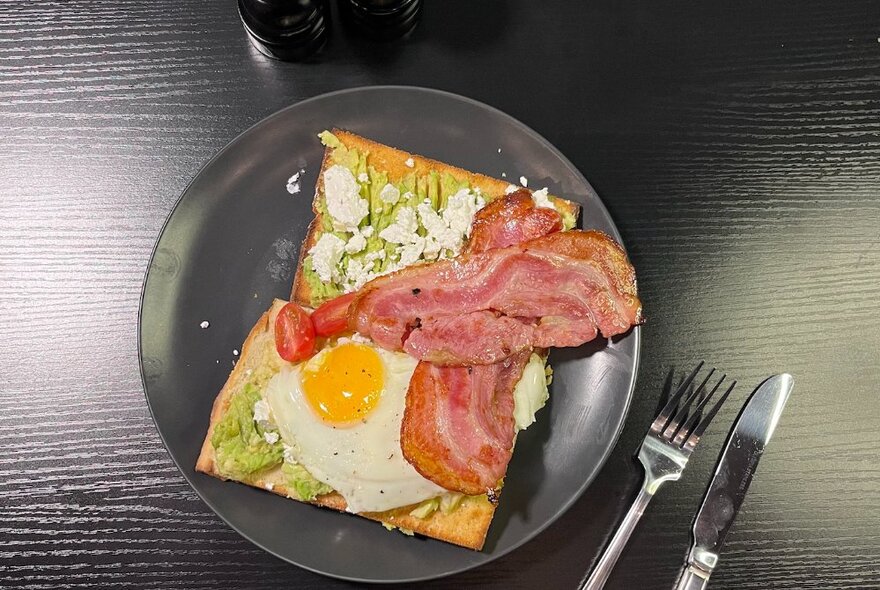 Toast squares with avocado, fried egg, feta and bacon strips on a black plate.