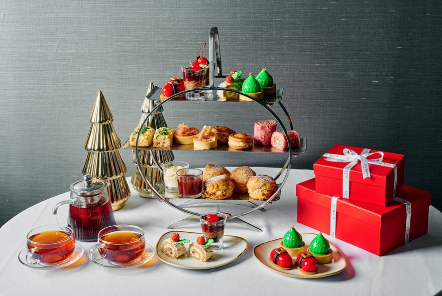 A festive-themed high tea on a round table, with a three tier stand of small items of food, a small decorative Christmas tree, and two red gift boxes.