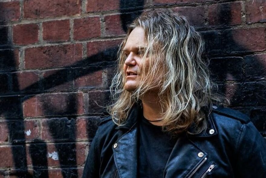 A man with wavy long hair wearing a leather jacket, standing against a red brick wall with black graffiti.