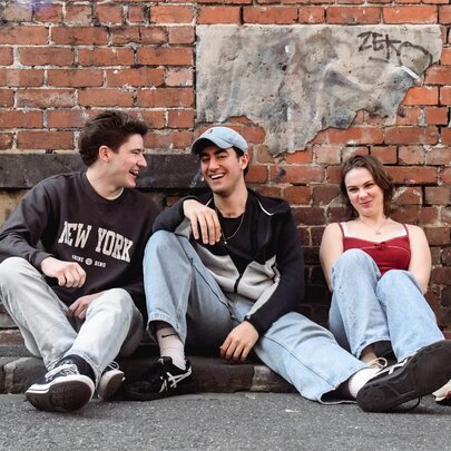 Three young people sitting casually in a gutter against an old brick building, relaxed and enjoying a laugh.