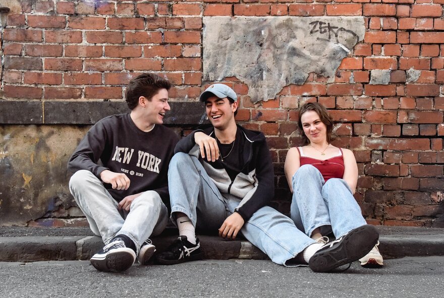 Three young people sitting casually in a gutter against an old brick building, relaxed and enjoying a laugh.