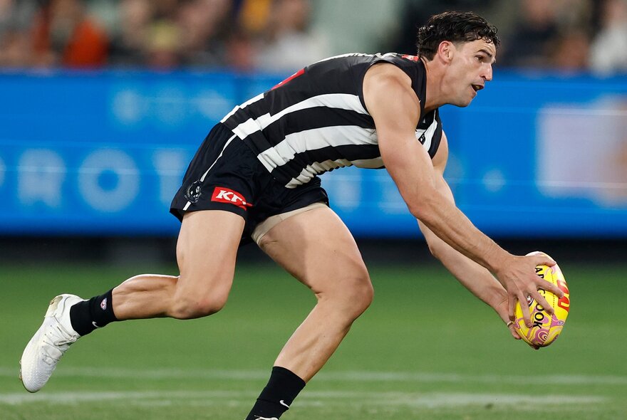 A Collingwood AFL football player reaching down to catch the football during a match.