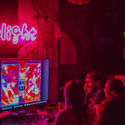 People seated and interacting with a screen monitor under a pink 'Delight' neon sign at an indoor bar venue.