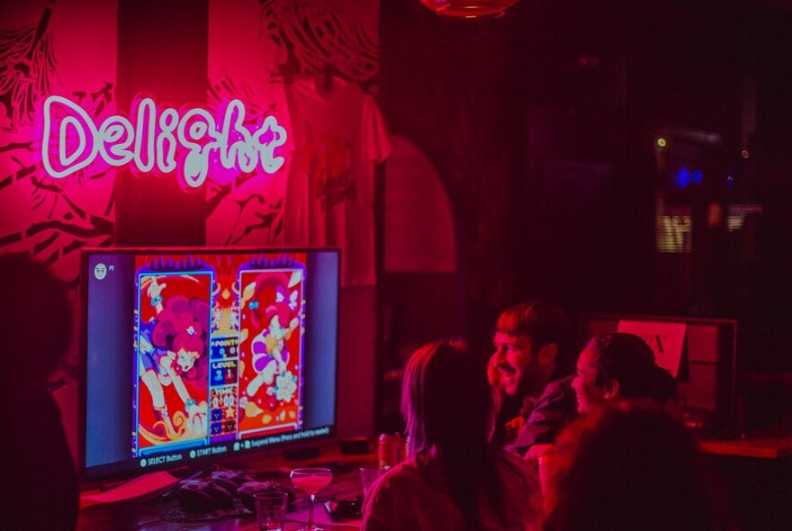 People seated and interacting with a screen monitor under a pink 'Delight' neon sign at an indoor bar venue.