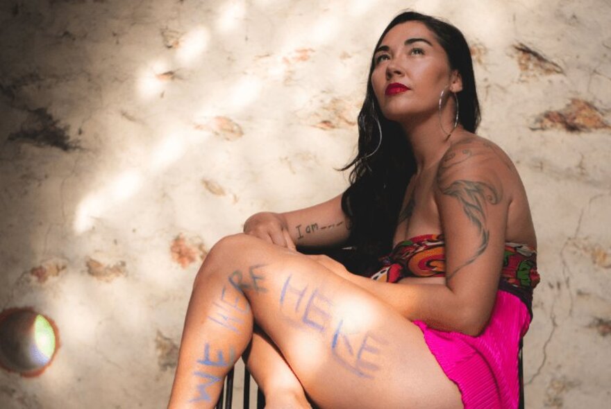 A female musician, Alphamama, posing in a brief pink outfit, with writing on her leg and tattoos on her arm.