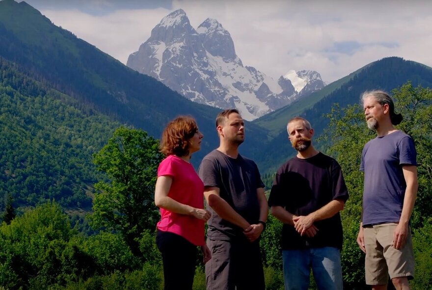 Four people standing on a green mountainside with snowcapped alps in the background.