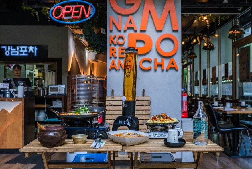 Modern Korean restaurant with industrial-style decor, and a long wooden table in the foreground laden with various dishes, and signage in rear on a pillar that reads 'Gangnam Pocha'.