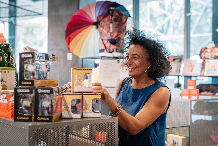 A woman in a shop laughing and reaching for one of the stacked boxes on display, a colourful umbrella in the background. 