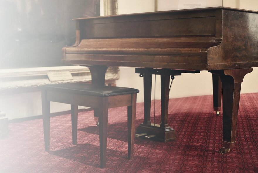 A flared photo showing a grand piano and matching piano stool, in a red carpeted room with cream coloured walls. 