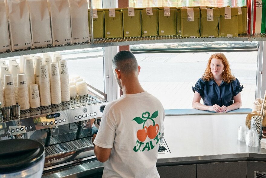 Interior of the St ALi coffee kiosk at University of Melbourne showing a person making a coffee at a machine, while a person waits on the other side of the takeaway window outside.