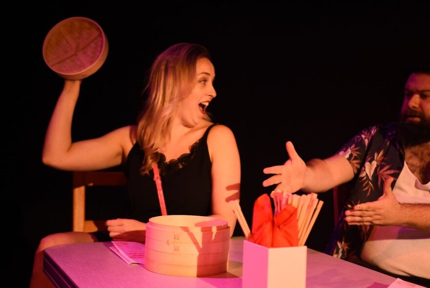 Two actors on stage performing, seated at a kitchen table and singing. 