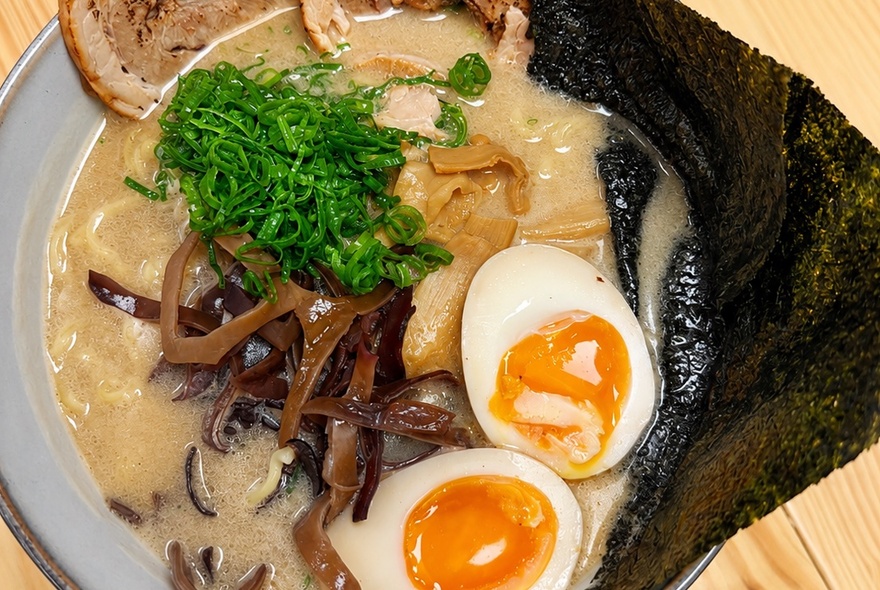 A bowl of tonkotsu ramen, featuring a creamy, broth topped with pork, a soft-boiled egg with a jammy yolk, mushrooms, and nori seaweed.