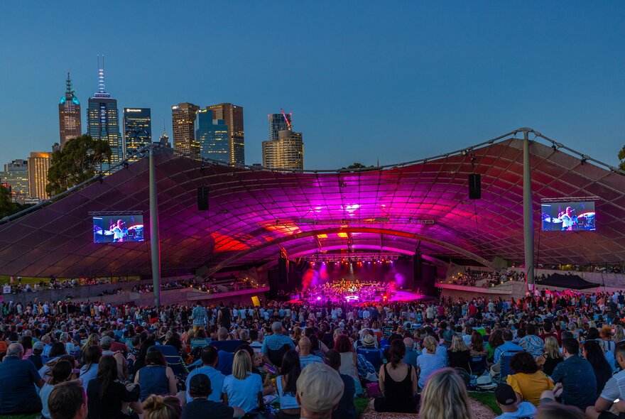 An crowd of people are watching an outdoor concert at dusk at Sidney Myer Music Bowl