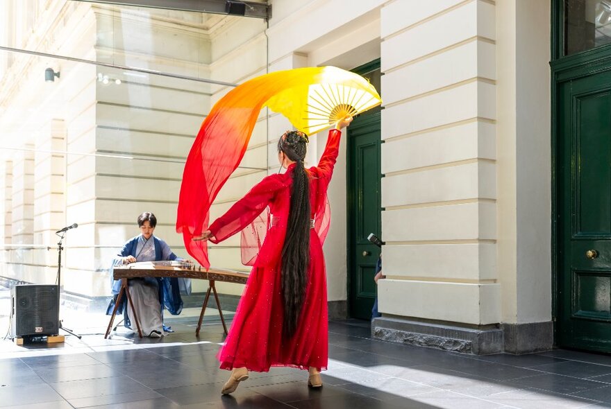 A woman performing a dance with a scarf in red and yellow tomes in a courtyard while another woman is seated playing an instrument. 