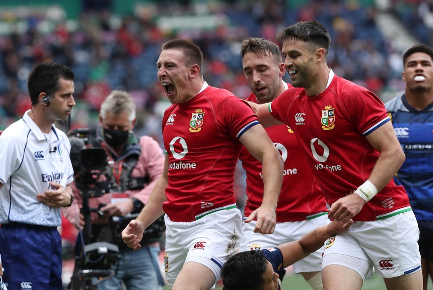 Rugby players from the Lions in red and white, on the sports field with a photographer close by, cheering and looking jubilant.