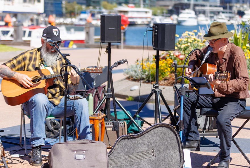 Two seated men playing guitars with small speakers behind them at an outdoor market.
