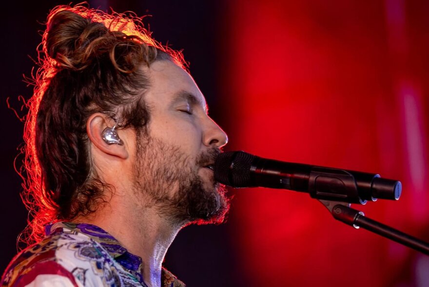 Profile of the head of singer Jeremy Loops, his hair is piled on top of his head in a loose bun, his eyes are closed and he sings into a microphone.