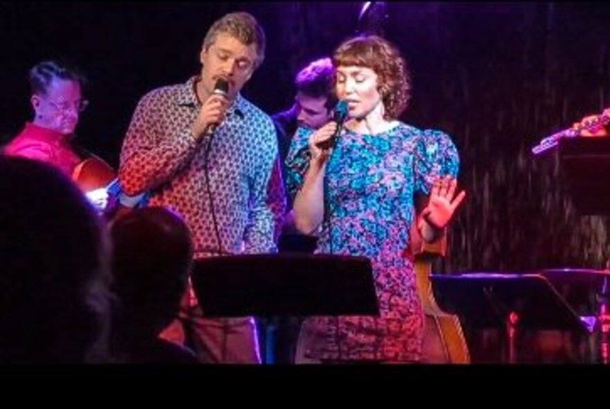 Male and female performers singing a duet with microphones on stage in front of seated audience.