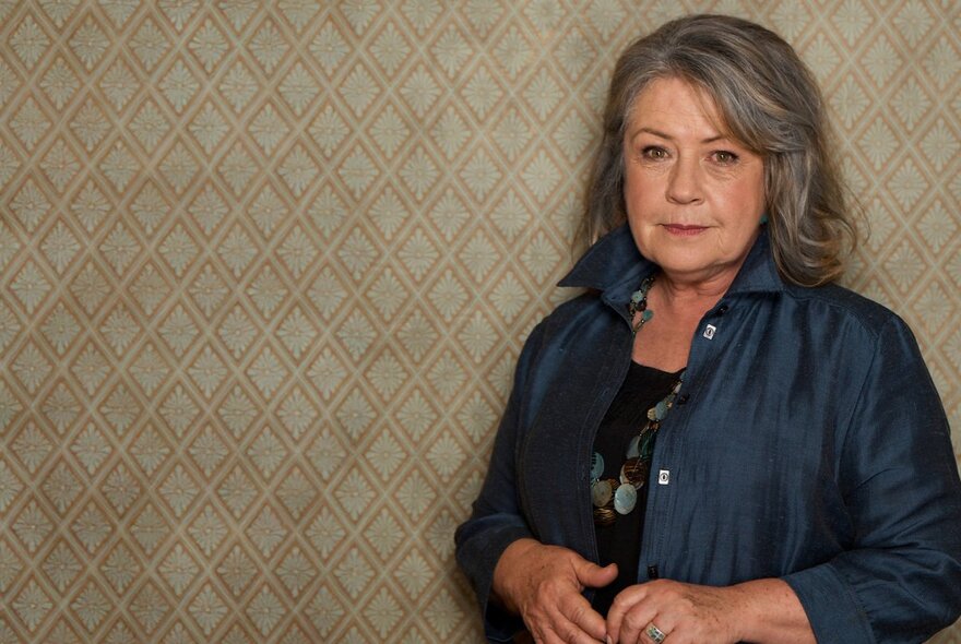 Actor Noni Hazelhurst, with a slight smile and her hands together at her waist, arrange to the right of the picture against a wall with a diamond-shaped beige pattern.