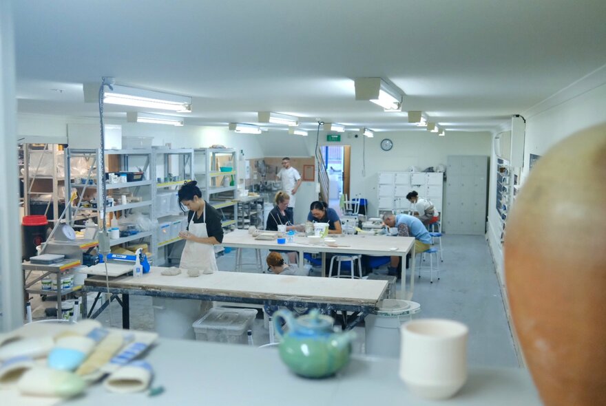 A brightly lit pottery studio with people creating at long tables and various pottery items around the room.