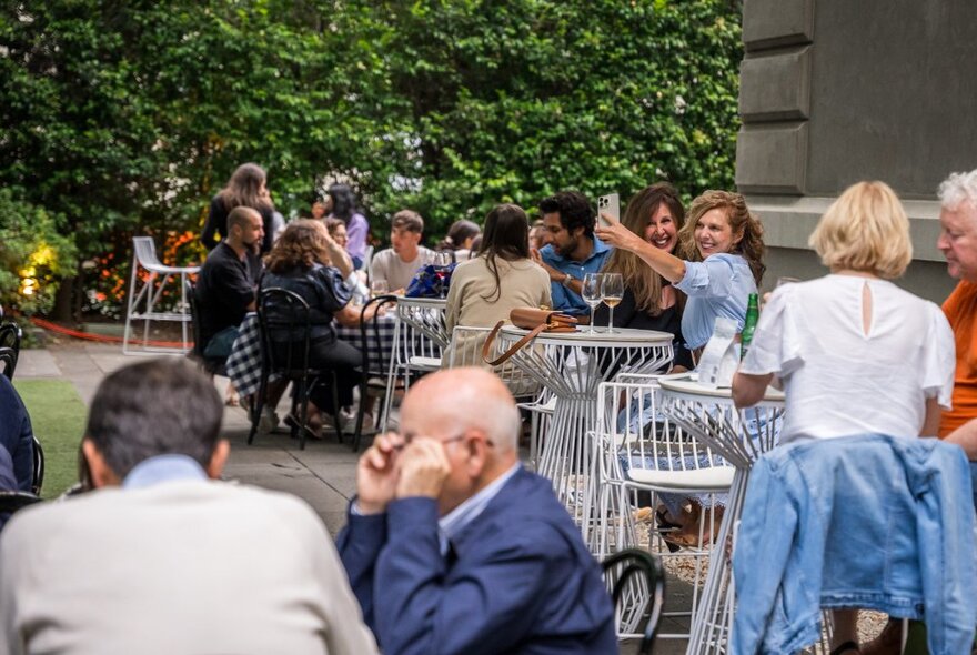 Diners seated at outdoor tables under trees, two women cheering with glasses.