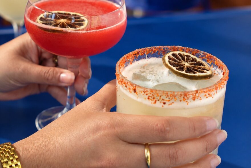 Two cocktails, one orange in colour and one pale with an orange rimmed-glass, both with dried citrus garnish. 
