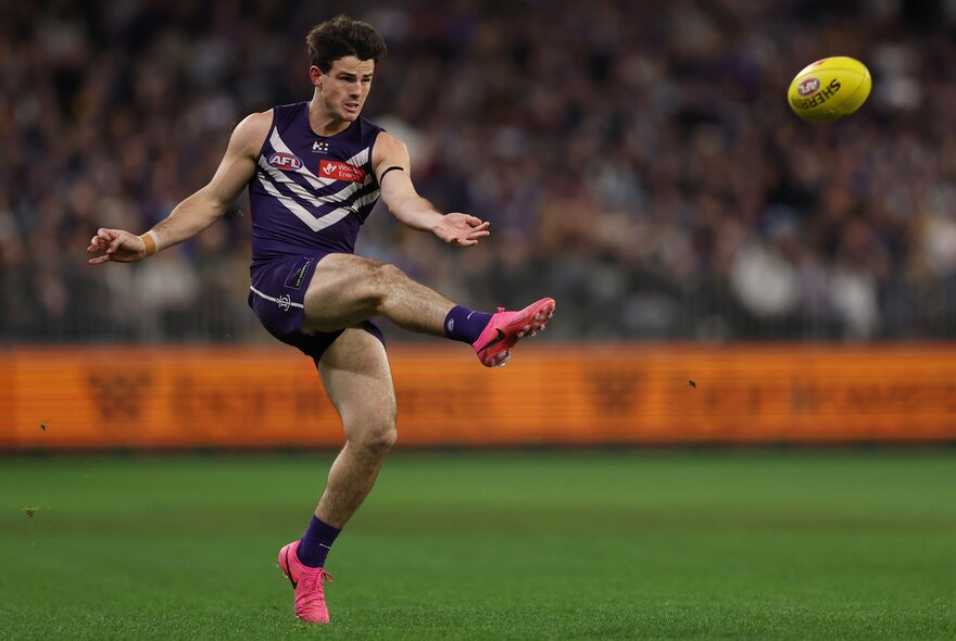 A Fremantle Dockers AFL player kicking a yellow ball during a game.