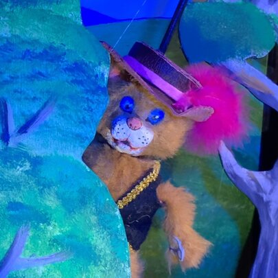 Puss in Boots cat puppet figure wearing a waistcoat and feathered hat, hiding behind illustrations of trees.