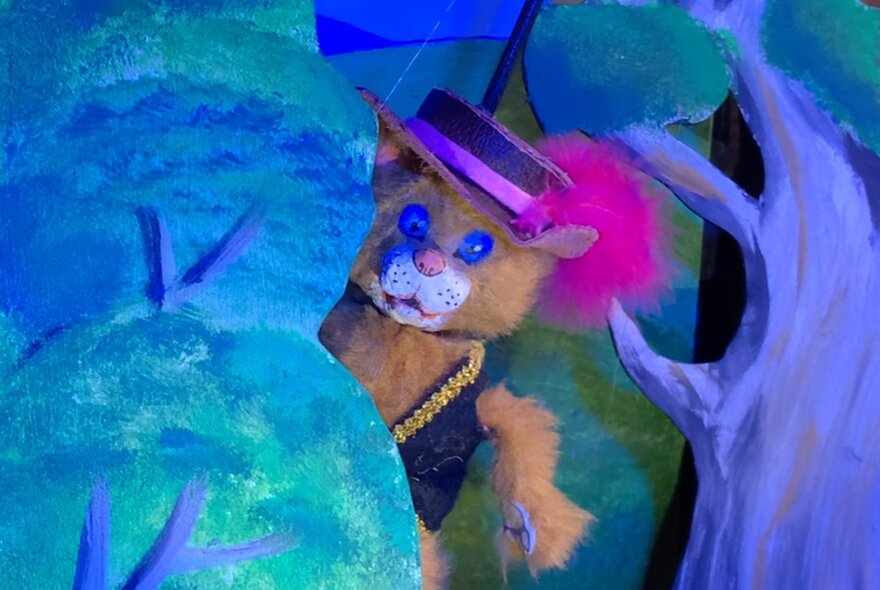 Puss in Boots cat puppet figure wearing a waistcoat and feathered hat, hiding behind illustrations of trees.