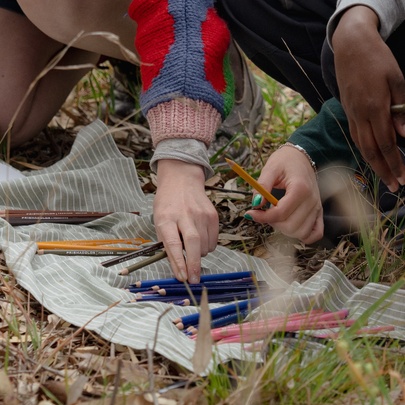 Hands reaching for pencils on a piece of fabric on leafy ground.