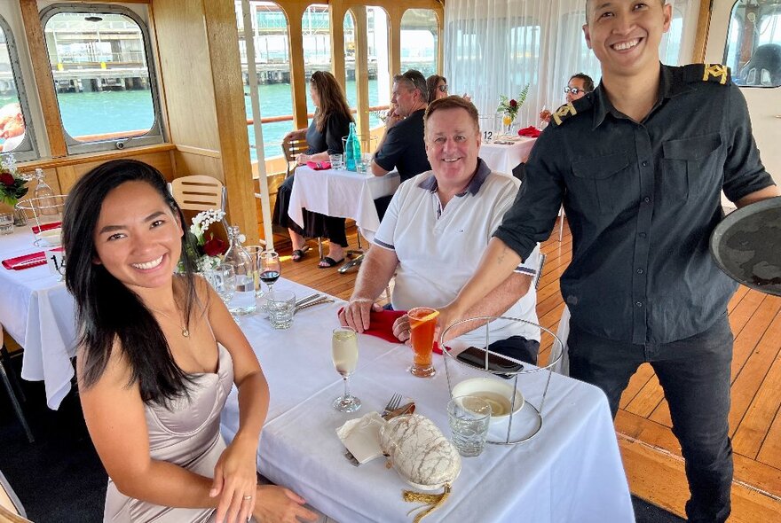 A smiling waiter serving drinks to a couple seated at a table on board the Lady Cutler Showboat, other diners behind them.