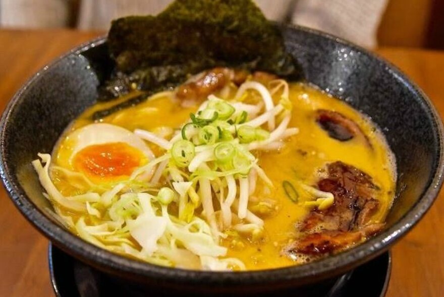 Ramen in a black bowl with egg, bean shoots and seaweed.