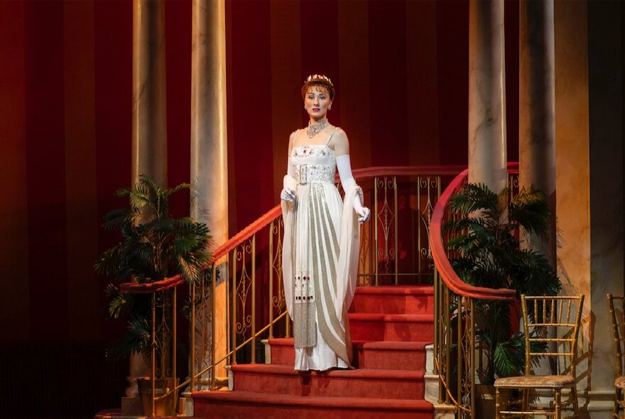 A scene from the musical My Fair Lady with Eliza Doolittle walking down a red velvet staircase wearing a beautiful strapless gown and tiara.