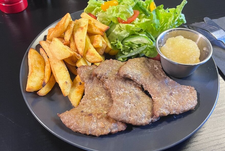 Minced pork shapes with chips, garden salad and apple sauce on a black plate.