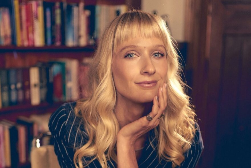 Dee Salmin, a smiling woman with wavy blonde hair resting her chin in her left hand, with a blurred wooden bookshelf filled with books behind her.