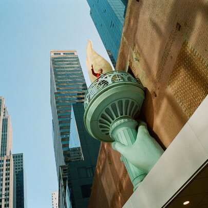 Low-angle, eye-level outdoor shot displays a sculpture replica of the Statue of Liberty's hand holding the green torch against a brown brick building facade on the right side of the frame, with tall skyscrapers against a blue sky in the background.