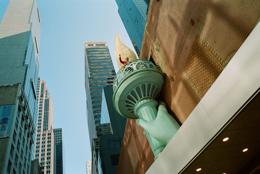 Low-angle, eye-level outdoor shot displays a sculpture replica of the Statue of Liberty's hand holding the green torch against a brown brick building facade on the right side of the frame, with tall skyscrapers against a blue sky in the background.