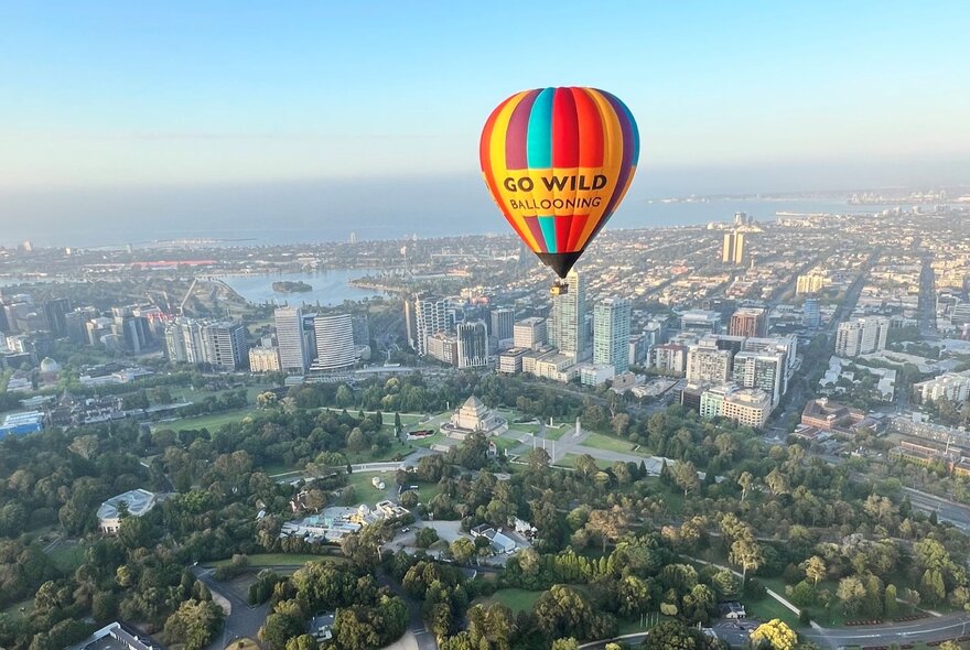 A hot air balloon in rainbow stripes with GO WILD Ballooning written on it floating high above Melbourne.