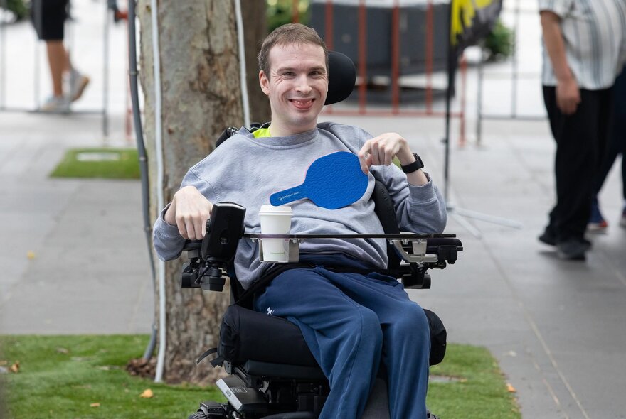A person smiling while seated in a motorized wheelchair and holding a table tennis bat, outdoors on a paved walkway.
